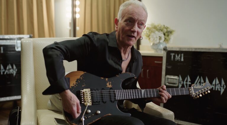 Jackson Phil Collen PC1-E: análisis de la nueva signature que sale del Custom Shop 10 Guitarra eléctrica Jackson Pro Series Signature Phil Collen PC1-E con acabado relic negro y herrajes dorados, sostenida por Phil Collen sentado en un camerino o zona de backstage durante una demostración del instrumento.