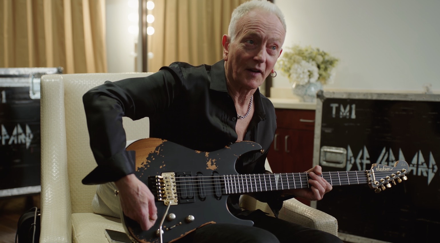 Jackson Phil Collen PC1-E: análisis de la nueva signature que sale del Custom Shop 1 Guitarra eléctrica Jackson Pro Series Signature Phil Collen PC1-E con acabado relic negro y herrajes dorados, sostenida por Phil Collen sentado en un camerino o zona de backstage durante una demostración del instrumento.