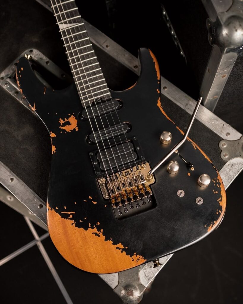 Jackson Phil Collen PC1-E: análisis de la nueva signature que sale del Custom Shop 5 Detalle de la guitarra eléctrica Jackson Phil Collen PC1-E con acabado relic negro desgastado, puente Floyd Rose dorado, configuración de pastillas HSS y controles metálicos sobre una superficie de transporte profesional.