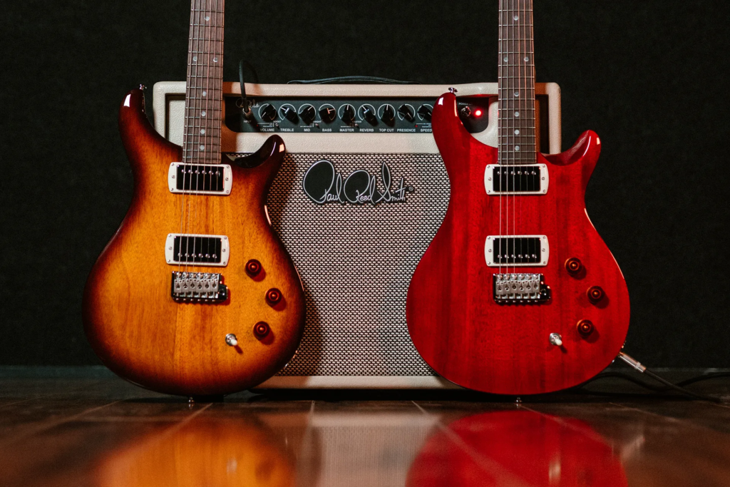 PRS DGT 15 Combo: análisis del combo de válvulas de 15 vatios de David Grissom 2 Amplificador PRS DGT 15 Combo situado entre dos guitarras eléctricas PRS con acabados sunburst y rojo translúcido, mostrando el panel de control frontal y la estética clásica del equipo en un entorno de demostración de guitarra.