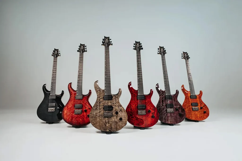 Colección de guitarras eléctricas de la gama PRS Tekton, mostrando varios modelos con tapas de arce figurado en acabados negro, rojo, natural, morado y naranja, equipados con configuración de doble humbucker, puente de trémolo y diseño de doble cutaway orientado a estilos modernos y de alto rendimiento.