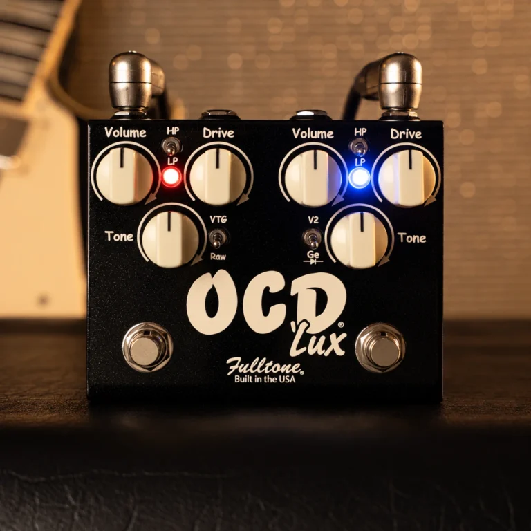 Pedal de overdrive Fulltone OCD'Lux First Edition con doble circuito OCD independiente, controles de volumen, drive y tono para cada canal, indicadores LED rojo y azul, y conmutadores de voicing dedicados, presentado sobre una superficie de estudio con un amplificador de guitarra desenfocado al fondo.