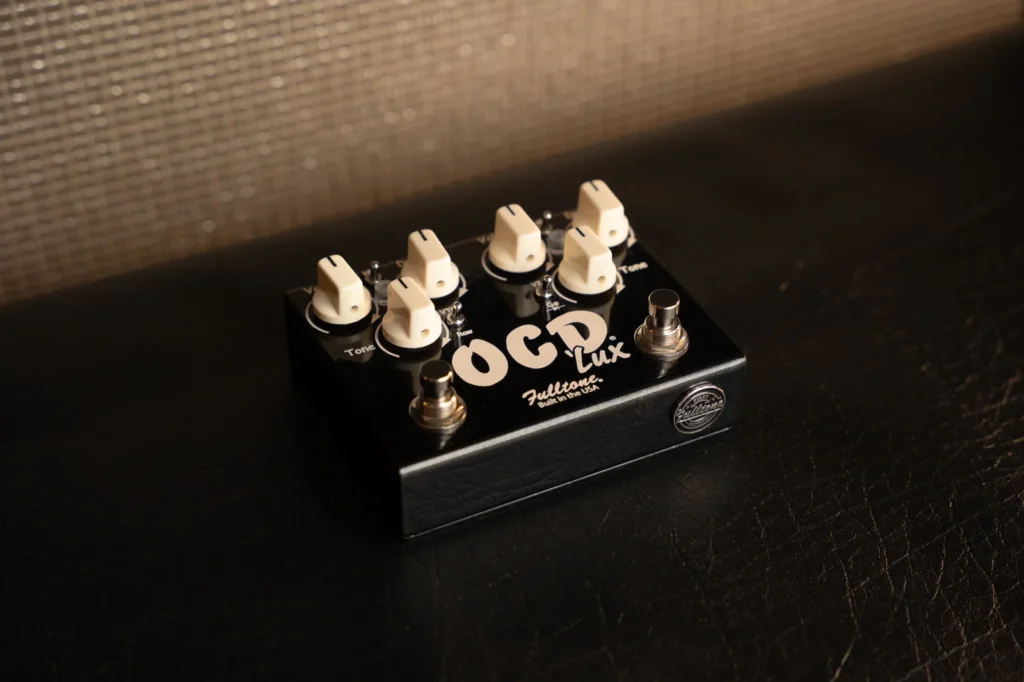 Pedal de overdrive Fulltone OCD'Lux First Edition fotografiado en perspectiva sobre una superficie oscura, mostrando su diseño de doble canal OCD, dos footswitches independientes, seis controles principales y el distintivo conmemorativo “First Edition” integrado en la carcasa negra.
