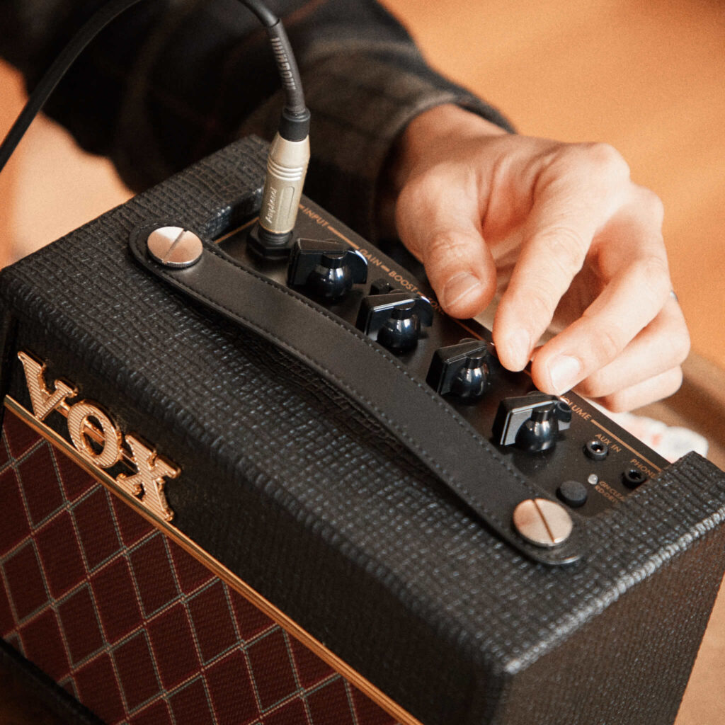 Vox AC Mini: análisis del mini ampli de escritorio de 10 W (¿ampli o juguete?) 7 Detalle del panel de control superior del amplificador portátil Vox AC Mini, con una mano ajustando los controles de ganancia, tono y volumen, mientras una guitarra eléctrica conectada al equipo aparece parcialmente visible al fondo.