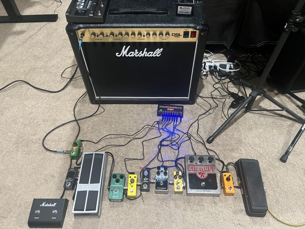 Configuración de guitarra eléctrica con amplificador Marshall DSL40C y cadena de pedales conectada por la entrada frontal, incluyendo wah, fuzz, overdrive, delay y afinador, distribuidos sobre una alfombra sin utilizar el bucle de efectos del amplificador.