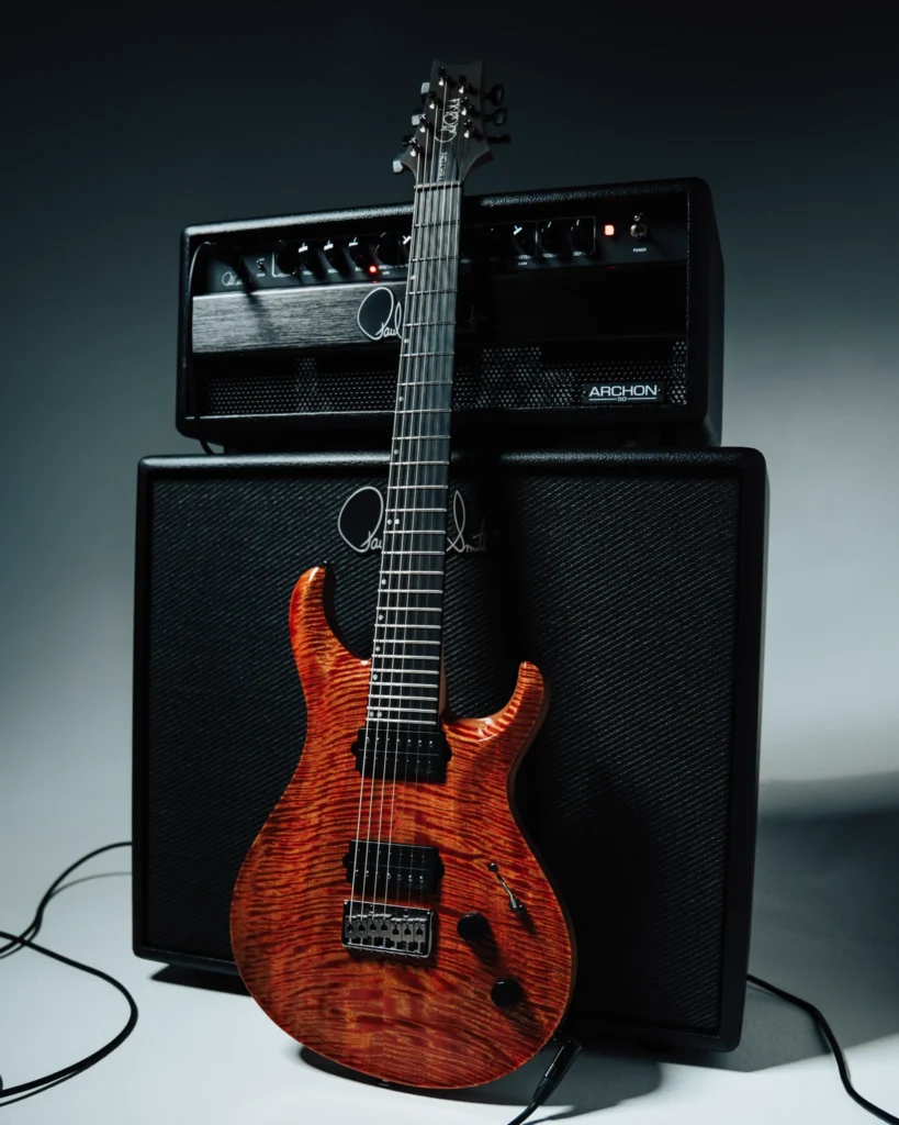 Guitarra de la gama PRS Tekton SVN de siete cuerdas con acabado naranja translúcido sobre tapa de arce figurado, apoyada frente a un cabezal PRS Archon 50 y una pantalla a juego, mostrando una configuración orientada a afinaciones extendidas, alta ganancia y estilos modernos de metal y rock progresivo.