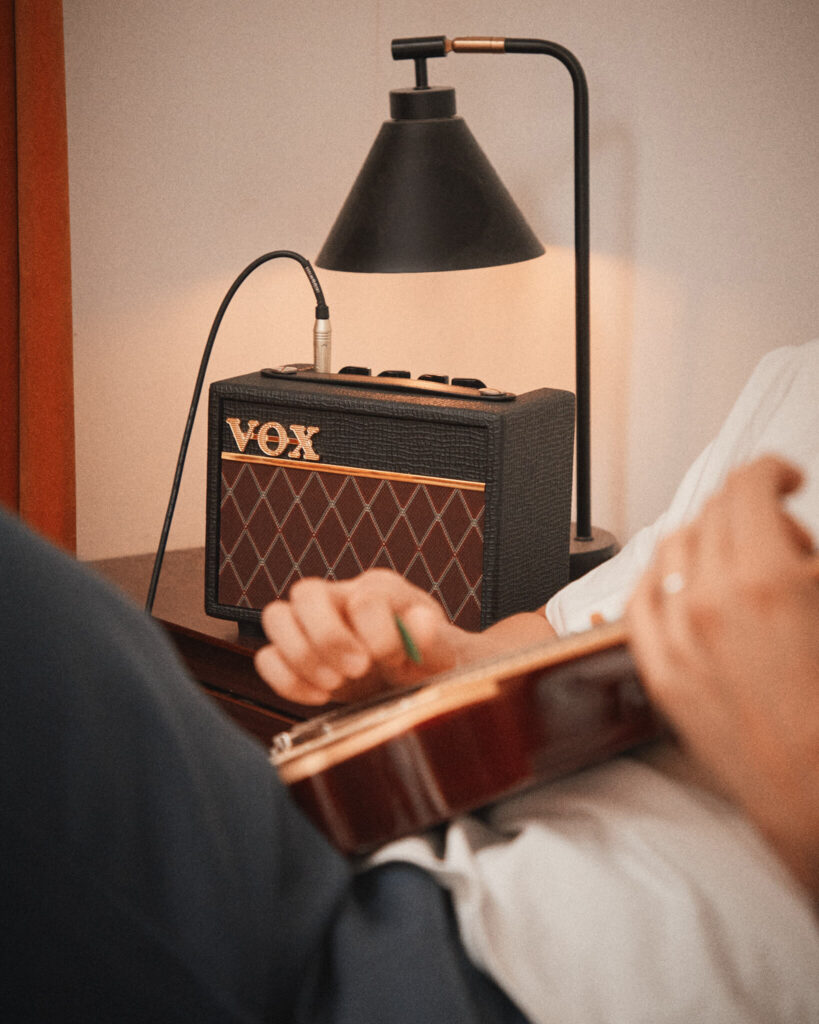 Vox AC Mini: análisis del mini ampli de escritorio de 10 W (¿ampli o juguete?) 4 Amplificador portátil Vox AC Mini colocado sobre una mesilla junto a una lámpara de escritorio, conectado a una guitarra eléctrica fuera de foco en primer plano, mostrando un entorno doméstico de práctica silenciosa y uso personal.